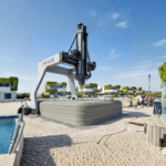 A robotic 3D printer arm layering concrete on a luxury villa in Dubai South, with completed futuristic white villas in the background under a blue sky.