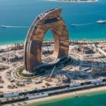 A panoramic view of Al Marjan Island in 2026, showing the nearly complete Wynn Resort tower dominating the skyline, surrounded by new luxury construction cranes and sparkling blue water.
